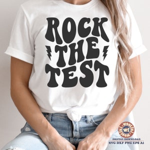 May include: White t-shirt with black text that reads "Rock the Test" in a retro font style. The text is stylized with lightning bolts on either side of the word "the".