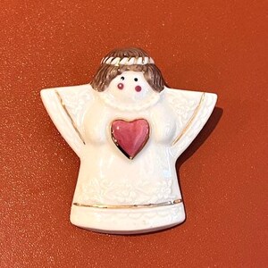 May include: A white ceramic angel figurine with a pink heart on the chest. The angel has brown hair, a gold halo, and gold trim on the wings and base. The figurine is on a reddish-orange surface.