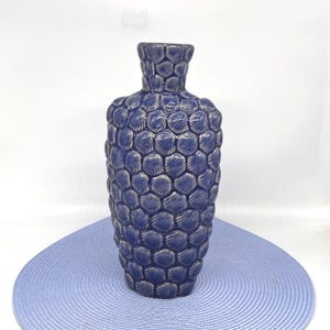 May include: A navy blue ceramic vase with a textured, shell-like pattern. The vase has a wide body and a narrow neck, standing on a light blue mat. The vase is approximately 10 inches tall.