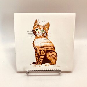 May include: A white ceramic tile with a hand-painted illustration of a brown and white tabby cat sitting and looking to the left. The cat has green eyes and a white patch on its chest. The tile is signed "Mary Mead" in the bottom right corner.