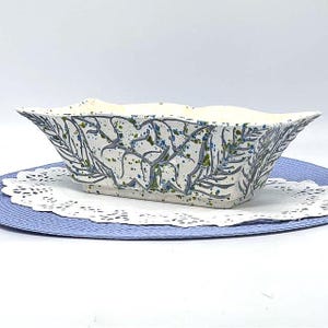 May include: A rectangular ceramic planter with a white base, adorned with a blue and green leaf pattern. The planter has a slightly flared shape and sits on a white doily and a blue mat. The design is hand-painted, giving it a unique, artistic look.