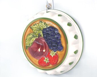 May include: A round, ceramic wall hanging with a fruit design. The center features a plum, grapes, and strawberries. The border is white with green accents and a gold-colored rim. A metal wire is attached for hanging.
