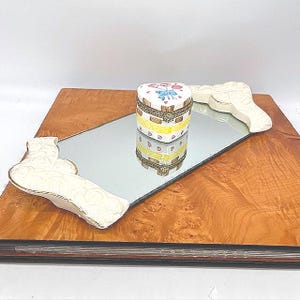 May include: A heart-shaped trinket box with a floral design sits on a mirrored tray with decorative, off-white handles. The box has a white base with yellow and pink accents, and a gold-colored metal frame. The tray rests on a wooden surface.
