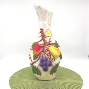 May include: A tall, off-white vase with a textured surface and a unique, asymmetrical top. The vase is adorned with colorful, raised relief fruit and vine details, including red apples, yellow pears, and purple grapes. The vase sits on a green placemat.