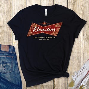 May include: Black t-shirt featuring a red and gold graphic with the text "Beasties" and "The King of Beats, New York, NY." The image includes blue jeans and white trainers on a wooden background.