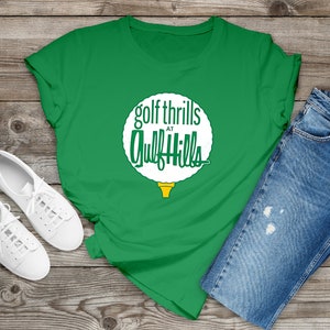 May include: A green t-shirt featuring a white graphic of a golf ball with the text "golf thrills at Gulf Hills". The shirt is laid out on a wooden surface with a pair of white trainers and blue jeans.