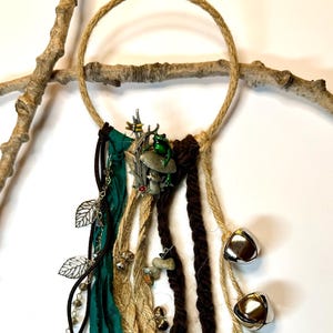 May include: A handcrafted dreamcatcher with a natural rope ring, adorned with green and brown fabric strips, silver bells, and decorative charms. The design includes a key, leaf charms, and a small frog figurine.