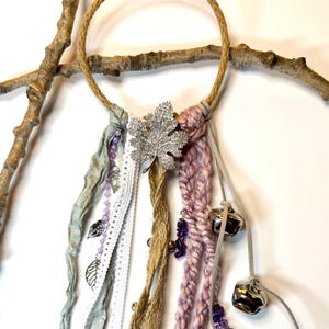May include: A decorative wall hanging featuring a woven rope circle adorned with fabric ribbons in shades of gray, white, and lavender. A silver leaf embellishment and small bells add detail. The piece is displayed on a natural branch.