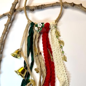 May include: A decorative wall hanging featuring a jute rope ring adorned with red, green, and cream-colored ribbons, lace, and small gold bells. Embellishments include a floral brooch and gold leaf charms. The piece is displayed on a natural branch.
