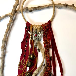May include: A handcrafted wall hanging featuring a woven rope ring adorned with burgundy ribbons, bells, and a metal charm. The charm is a winged figure with a heart. A vintage key hangs from the bottom, adding a rustic touch. The piece is suspended from a natural branch.