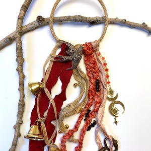 May include: A decorative wall hanging featuring a jute rope circle, adorned with red fabric, gold bells, and a metal bird. It includes strands of braided yarn, beads, and charms, including a key and crescent moon. The piece is attached to a tree branch.