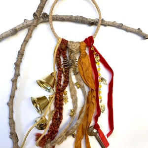May include: A handcrafted wall hanging featuring a rope ring adorned with various textures and embellishments. It includes strands of braided yarn, fabric ribbons in shades of red and gold, brass bells, and decorative charms, including a key and a scorpion.