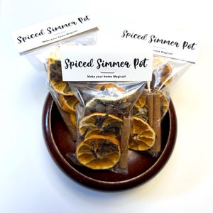 May include: A wooden bowl holds three clear plastic bags of spiced simmer pot ingredients. Each bag contains dried orange slices, cinnamon sticks, cloves, and star anise. The bags are labeled "Spiced Simmer Pot" with the tagline "Make your home Magical!"