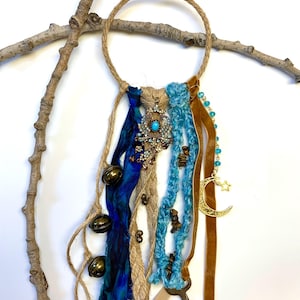 May include: A bohemian-style wall hanging featuring a jute rope ring with hanging ribbons in blue and brown. Embellishments include a vintage-style pendant with a turquoise stone, small bells, and a key. A crescent moon and star charm are also included.