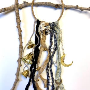 May include: A decorative wall hanging featuring a jute rope ring adorned with various embellishments. Includes brass bells, black and tan fabric strands, and a gold crescent moon charm. The hanging is suspended from a natural branch.
