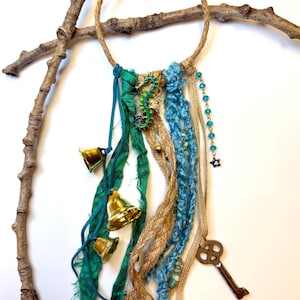 May include: A handcrafted dream catcher with a jute rope ring, adorned with teal fabric strips, gold bells, and blue yarn. A seahorse charm and a vintage key add whimsical detail. The dream catcher is displayed on a white background.