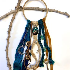 May include: A handcrafted dreamcatcher featuring a woven jute ring, adorned with teal and brown ribbons, a silver crescent moon charm, and a pendant with a miniature landscape scene. Additional embellishments include bells, beads, and a vintage key.