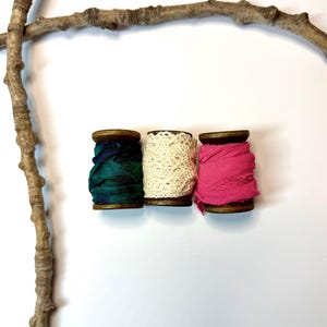 May include: Three wooden spools with colorful fabric ribbon: teal, cream lace, and hot pink. A natural branch curves across the top and left side of the image, set against a white background. Ideal for crafting, sewing, or decorative projects.