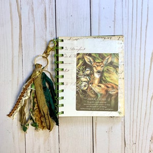 May include: A green and gold tassel hangs from a small, spiral-bound journal with a white cover. The journal features a printed image of a deer and the text "DEER LOVE. Be gentle with yourself. You heal with the power of love. See the light in all things. Acknowledge your self worth and grace."