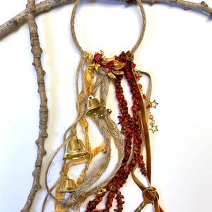 May include: A handcrafted wall hanging featuring a rope circle adorned with gold bells, red and gold floral accents, and strands of fabric in various textures and colors. Decorative stars and a key charm add detail.