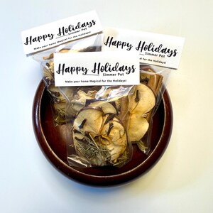 May include: A wooden bowl holds three clear bags of simmer pot ingredients. Each bag has a white label with the text "Happy Holidays" and "Make your home Magical for the Holidays!". The bags contain dried fruit and spices.