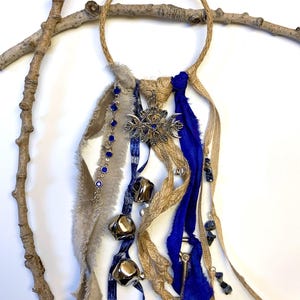 May include: A handcrafted wall hanging featuring a jute rope ring, adorned with blue and tan fabric strips, silver bells, and a silver compass pendant. The design includes blue gemstone accents and a small key charm, creating a bohemian aesthetic.