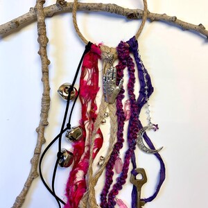May include: A decorative wall hanging featuring a jute ring adorned with pink, purple, and tan fabric strands. Silver charms include a hand, moon, and key, with dangling bells and crystals. The piece is displayed on a natural branch.