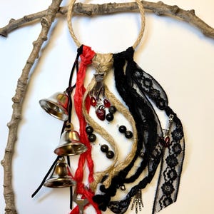 May include: A decorative wall hanging featuring a jute rope ring adorned with silver bells, black lace, red ribbons, and various charms, including a key and a heart. The design incorporates a rustic aesthetic with gothic elements.