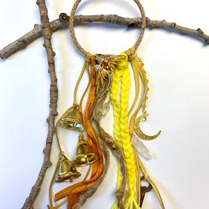 May include: A handcrafted dreamcatcher with a jute rope ring, adorned with yellow, orange, and brown ribbons, gold bells, and charms. The design includes a gold crescent moon, a key, and a crystal, creating a bohemian aesthetic.