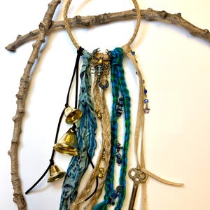 May include: A handcrafted dreamcatcher with a jute rope ring, adorned with blue and green fabric, brass bells, and a vintage key. A brass scorpion charm and blue beads add detail to this bohemian style wall hanging.