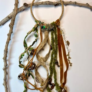 May include: A decorative wall hanging featuring a circular rope design with hanging embellishments. It includes brown suede ribbons, green faux moss, and small silver bells. A decorative insect and leaf charms add detail.