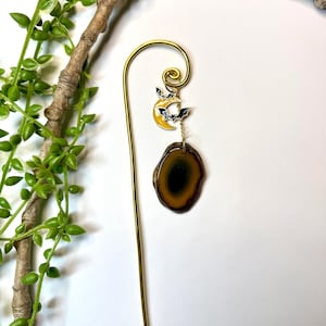 May include: A gold-colored metal bookmark with a Halloween theme. The top features a yellow crescent moon with black bats. A brown and green agate slice hangs below. Green leaves and a brown branch are in the background.