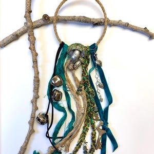 May include: A handcrafted wall hanging featuring a woven jute ring adorned with teal ribbons, silver bells, and decorative charms. The centerpiece includes a silver-toned embellishment with green accents and dangling keys.