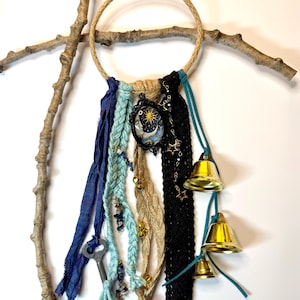 May include: A handmade dreamcatcher with a woven rope ring, adorned with blue and beige fabric strips, a key, and gold bells. A small pendant with a sun and moon design hangs from the center. The dreamcatcher is attached to a wooden branch.