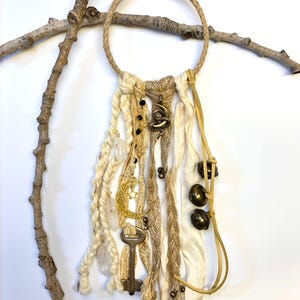 May include: A bohemian-style wall hanging featuring a jute rope ring with cream-colored fabric and yarn accents. Adorned with brass charms, including a crescent moon, key, and snake, along with small bells and clear crystals. The piece is displayed against a white background.