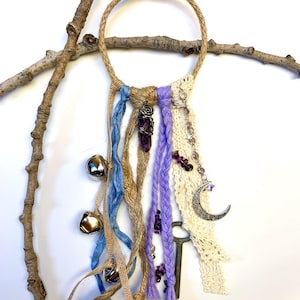 May include: A handcrafted dream catcher with a jute ring, adorned with blue and purple ribbons, silver bells, a crescent moon charm, and a key. The design incorporates a lace panel and amethyst beads, creating a bohemian aesthetic.