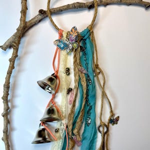 May include: A handcrafted dream catcher with a jute ring, adorned with silver bells, teal fabric, and butterfly charms. Features cream-colored lace, pink accents, and a natural branch for hanging. A unique, bohemian-style wall decor.