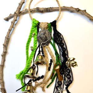 May include: A decorative wall hanging featuring a woven jute ring with green, black, and tan yarn accents. It includes a silver pendant, small bells, black beads, a key, and black lace. A twig is in the background.