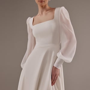 May include: A white wedding dress with a square neckline and long sleeves. The sleeves are made of a sheer fabric and have a puffed look. The dress has a fitted bodice and a flowing skirt.