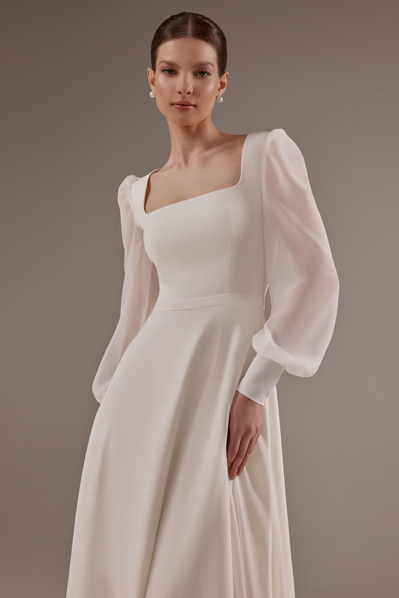 May include: A white wedding dress with a square neckline and long sleeves. The sleeves are made of a sheer fabric and have a puffed look. The dress has a fitted bodice and a flowing skirt.
