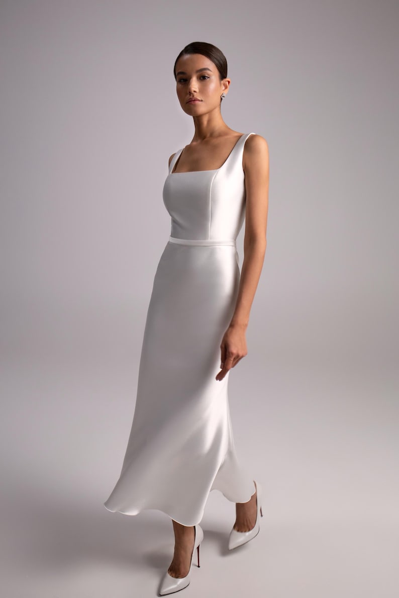 A white midi dress with a square neckline and a fitted bodice. The skirt is sheath and flows to the mid-calf. The dress is made of a smooth mikado, satin-like fabric. The dress features a thin, same-color waist band.