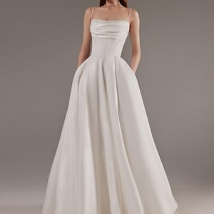 May include: A white wedding gown with a fitted bodice and a full, flowing skirt. The dress features a sweetheart neckline and thin spaghetti straps. The model is standing with hands in pockets, showcasing the dress's elegant design.