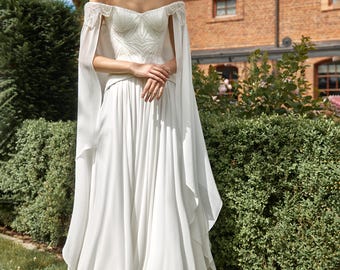 May include: A floor-length white off-the-shoulder gown featuring a structured bodice with intricate geometric embroidery, delicate beaded shoulder detailing, and flowing cape-style sleeves that drape down to the floor over a full, pleated skirt.