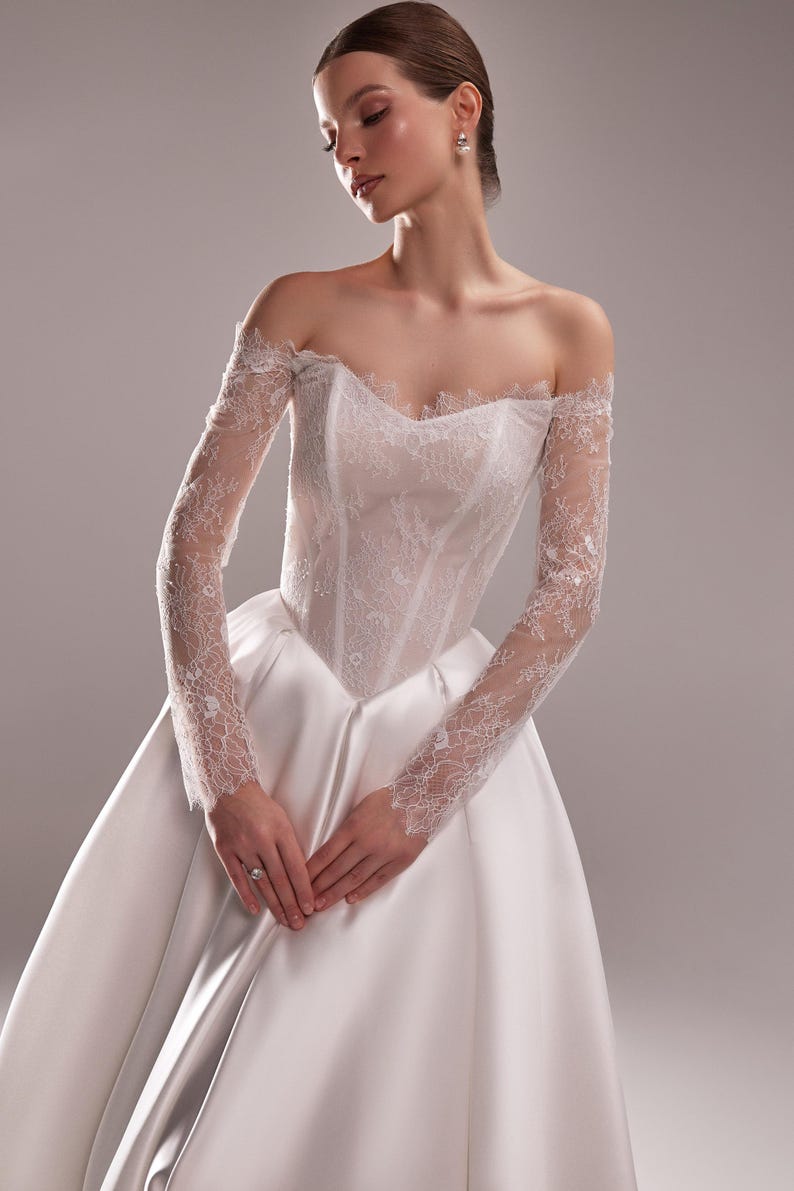 K&ouml;nnte beinhalten: Wei&szlig;es Brautkleid mit Satinrock und Spitzenoberteil. Die schulterfreien &Auml;rmel sind aus zarter Spitze gefertigt. Das Kleid hat ein klassisches, elegantes Design, perfekt f&uuml;r eine Hochzeit.
