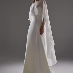 May include: A white bridal gown with a flowing skirt and a sheer cape overlay. The dress features a fitted bodice and a simple, elegant design. The cape drapes gracefully from the shoulders, adding a touch of sophistication.