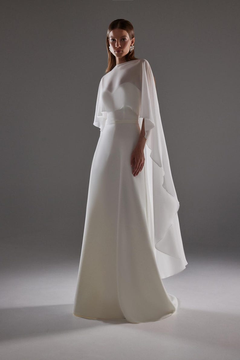 May include: A white bridal gown with a flowing skirt and a sheer cape overlay. The dress features a fitted bodice and a simple, elegant design. The cape drapes gracefully from the shoulders, adding a touch of sophistication.