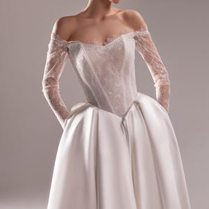 K&ouml;nnte beinhalten: Ein wei&szlig;es schulterfreies Hochzeitskleid. Das Oberteil ist aus Spitze und die langen &Auml;rmel sind transparent. Der Rock ist ein voller Ballkleid-Stil aus Satin. Das Model tr&auml;gt das Kleid mit den H&auml;nden in den Taschen.