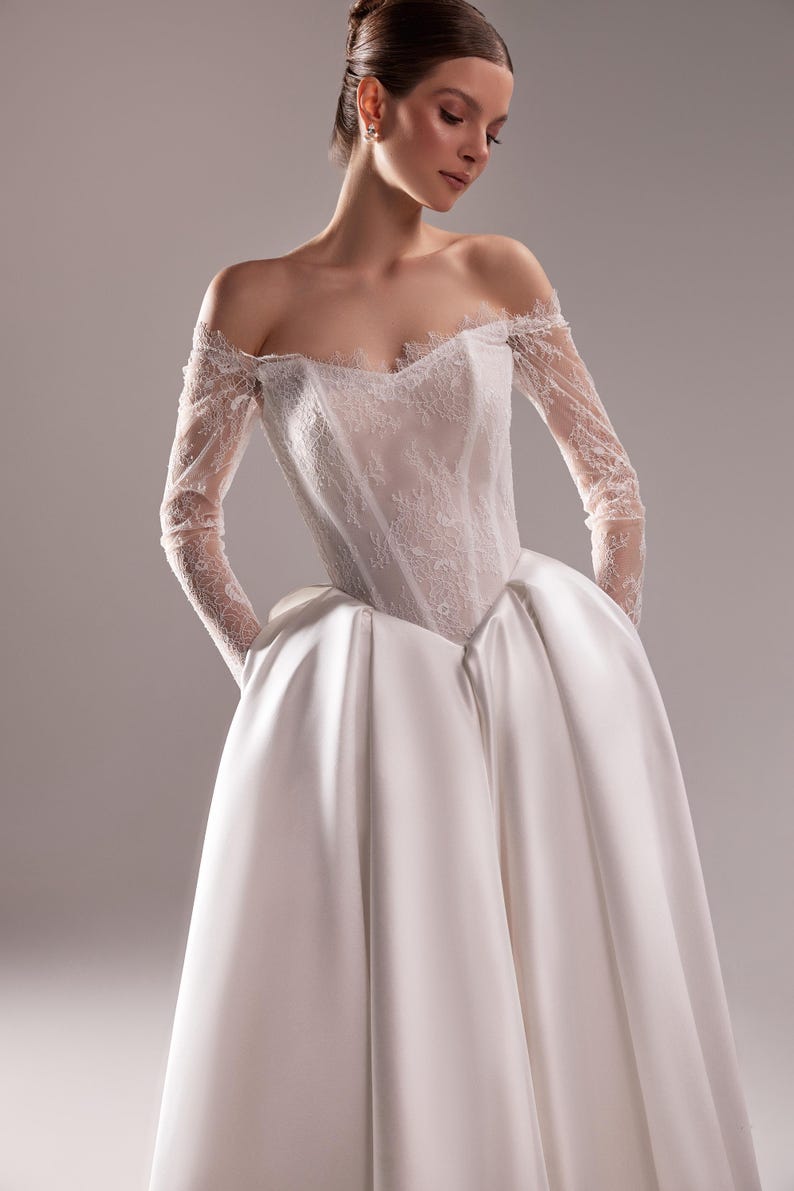 K&ouml;nnte beinhalten: Ein wei&szlig;es schulterfreies Hochzeitskleid. Das Oberteil ist aus Spitze und die langen &Auml;rmel sind transparent. Der Rock ist ein voller Ballkleid-Stil aus Satin. Das Model tr&auml;gt das Kleid mit den H&auml;nden in den Taschen.