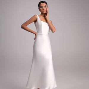 May include: A woman wearing a white sleeveless wedding dress with a fitted bodice and a flowing skirt. She is wearing white heels and has her hand on her cheek.