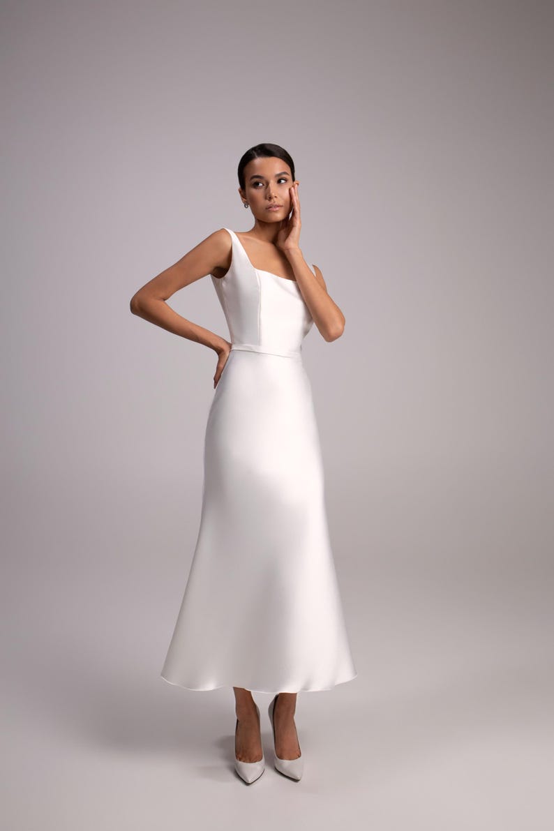 May include: A woman wearing a white sleeveless wedding dress with a fitted bodice and a flowing skirt. She is wearing white heels and has her hand on her cheek.